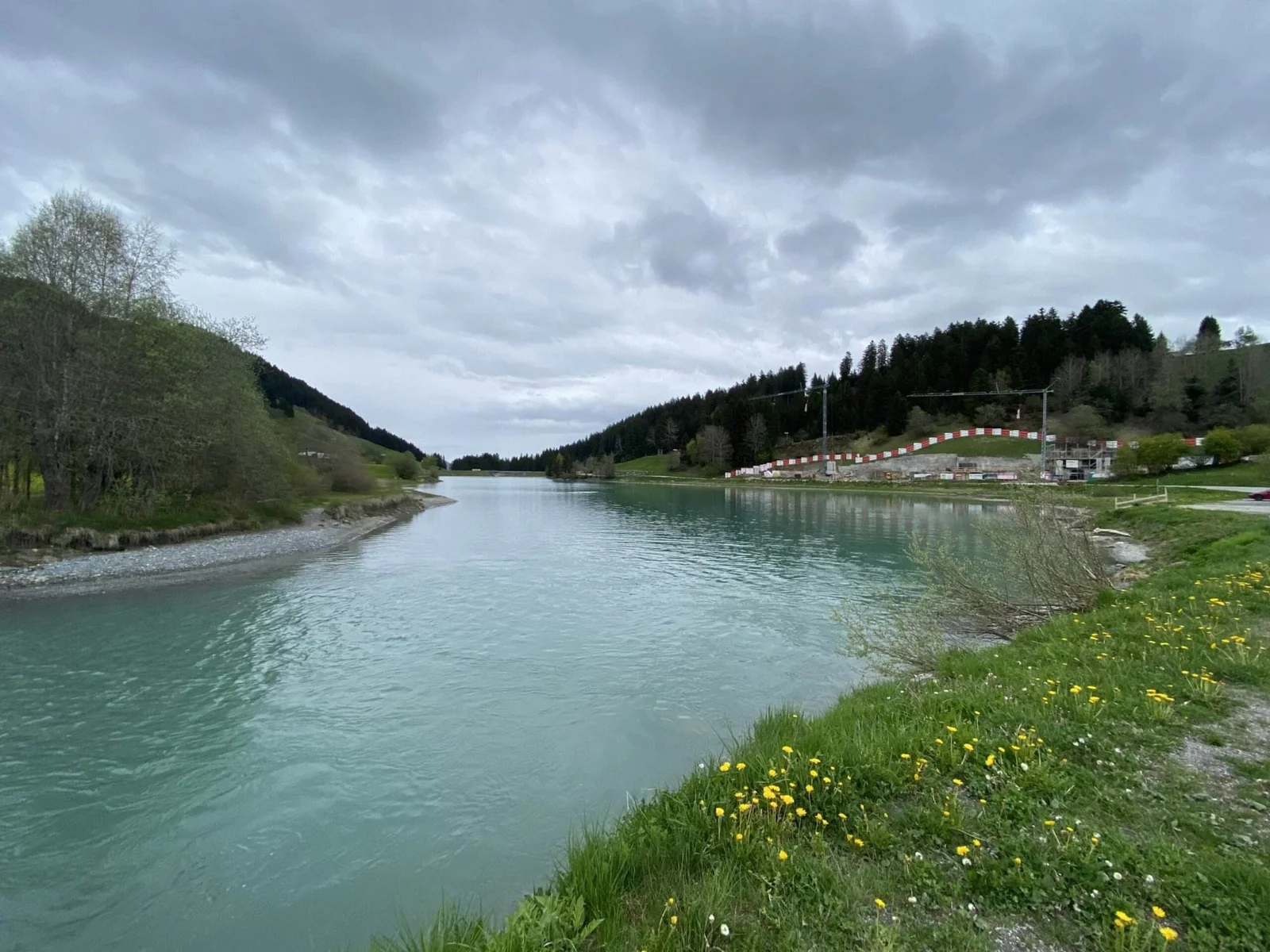 Türkisfarbener See mit sanft geschwungenem Ufer, umgeben von Wiesen und Wald, rechts eine kleine Baustelle mit rot-weißer Absperrung unter bewölktem Himmel.