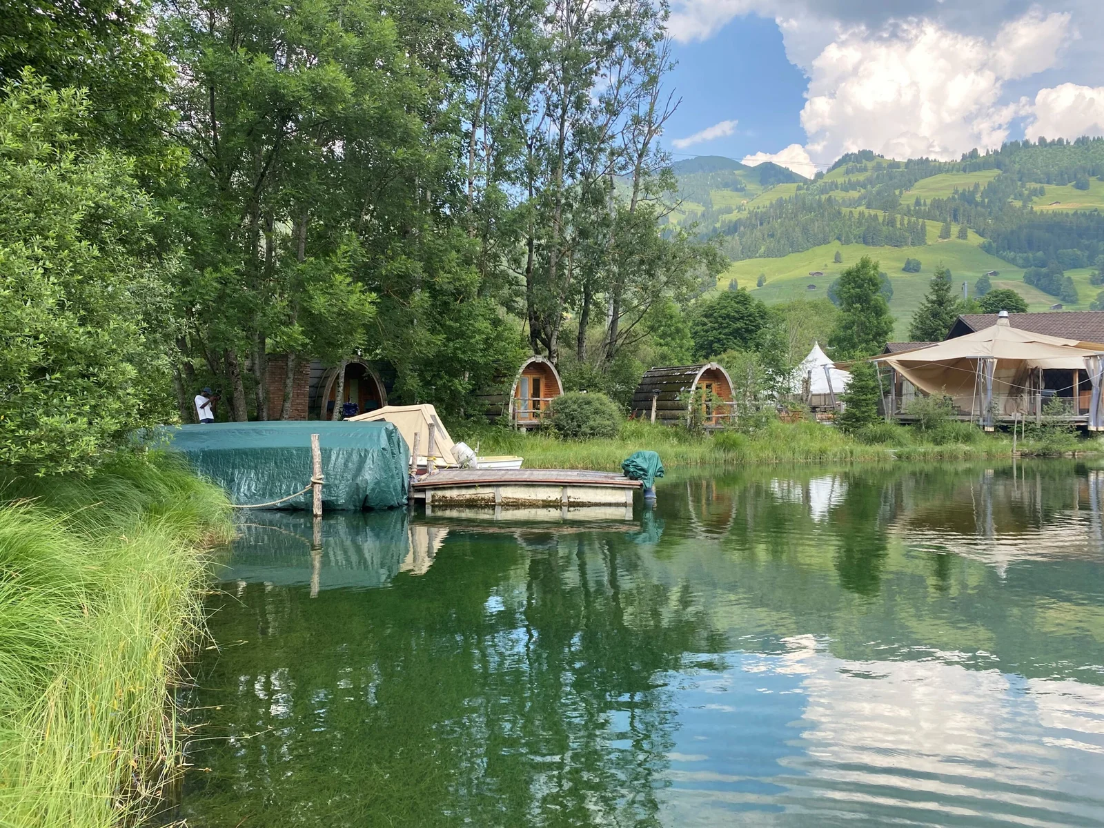 Kleiner See mit Steg, Hütten und grüner Berglandschaft.