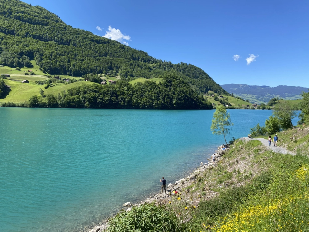 Türkisfarbener Bergsee mit klarem Wasser, umgeben von grünen Hügeln und bewaldeten Hängen. Am rechten Ufer verläuft ein schmaler Weg, auf dem mehrere Personen spazieren, während unten am Seeufer eine Person am Wasser steht. Im Hintergrund liegen verstreute Häuser in der Landschaft unter blauem Himmel.