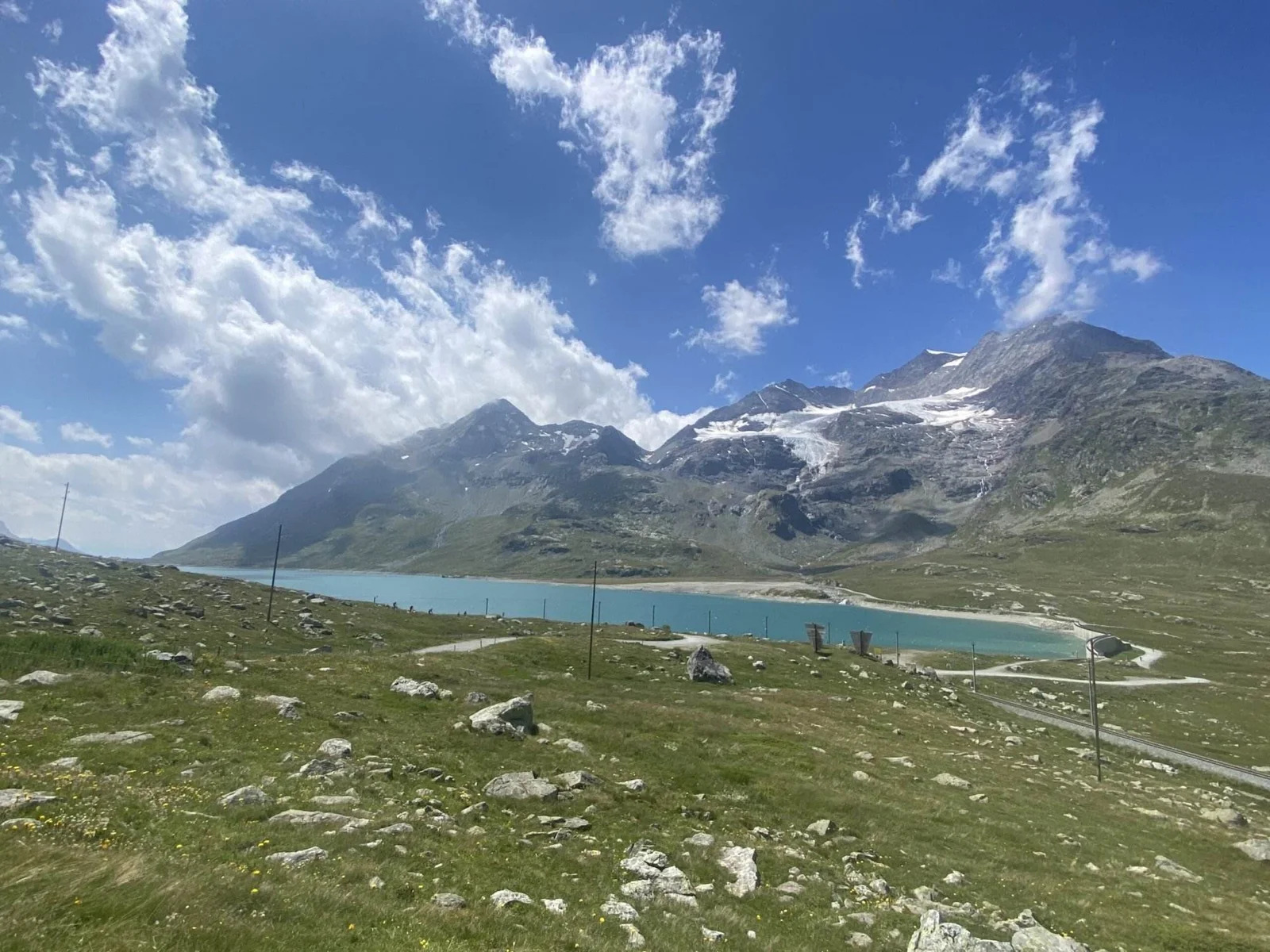Weite alpine Landschaft mit türkisfarbenem Bergsee, umgeben von grünen Wiesen und verstreuten Felsen. Im Hintergrund ragen hohe Berge mit Gletscherresten und Schneefeldern unter einem wolkigen, blauen Himmel auf.