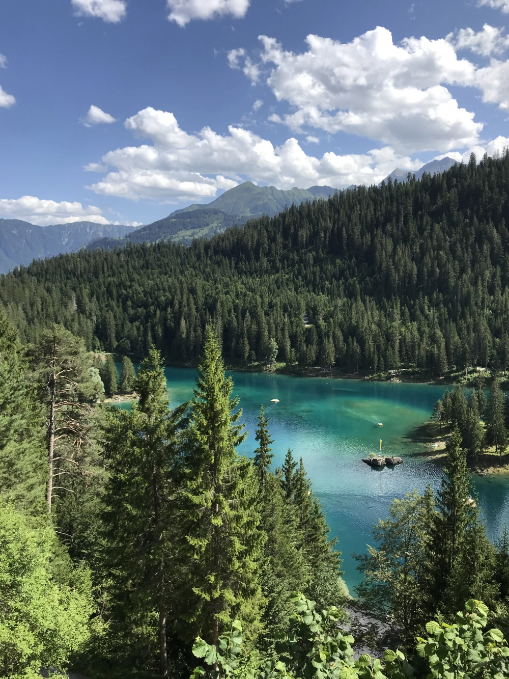 Türkisblauer Bergsee zwischen steilen, bewaldeten Hängen, unter blauem Himmel mit einzelnen weißen Wolken.