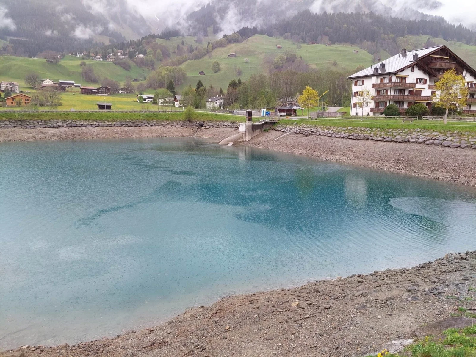 Türkisfarbener See mit steinigem Ufer, Wohnhäusern am Rand und grünen Hügeln im Hintergrund.