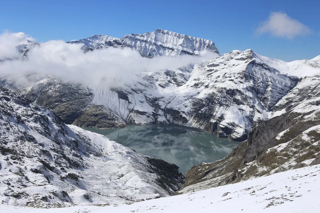 Lac d'Émosson: Verschneite Berge und ein ruhiger Bergsee unter blauem Himmel.