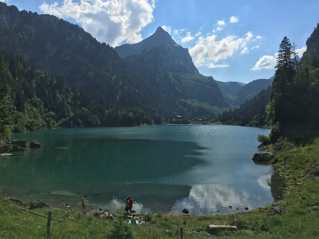 Idyllischer Lac de Tanay in den Bergen, mit Menschen am Ufer und Spiegelung der Wolken im Wasser.