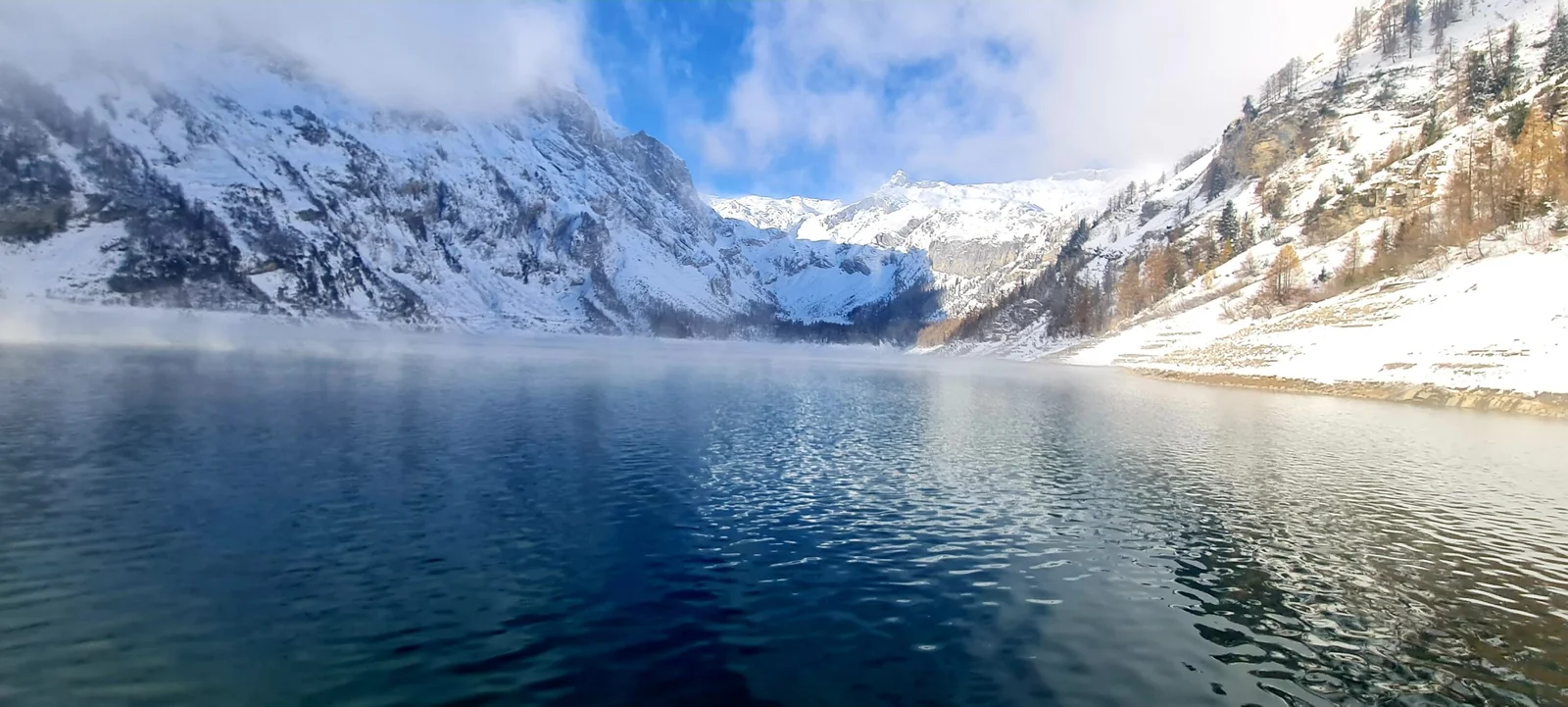 Winterlandschaft am Lac de Tseuzier: Verschneite Berge und ruhiger See unter blauem Himmel.