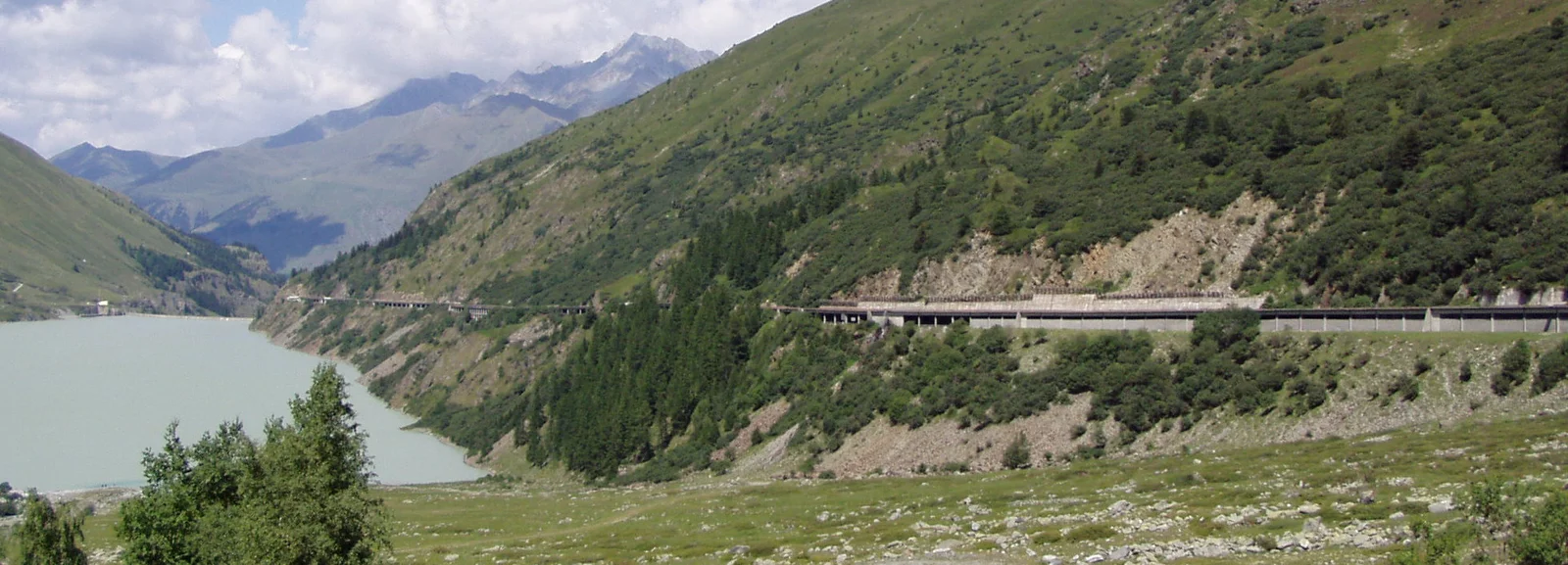 Lac des Toules mit Bergstraße und grünen Hängen. Bergpanorama im Hintergrund.