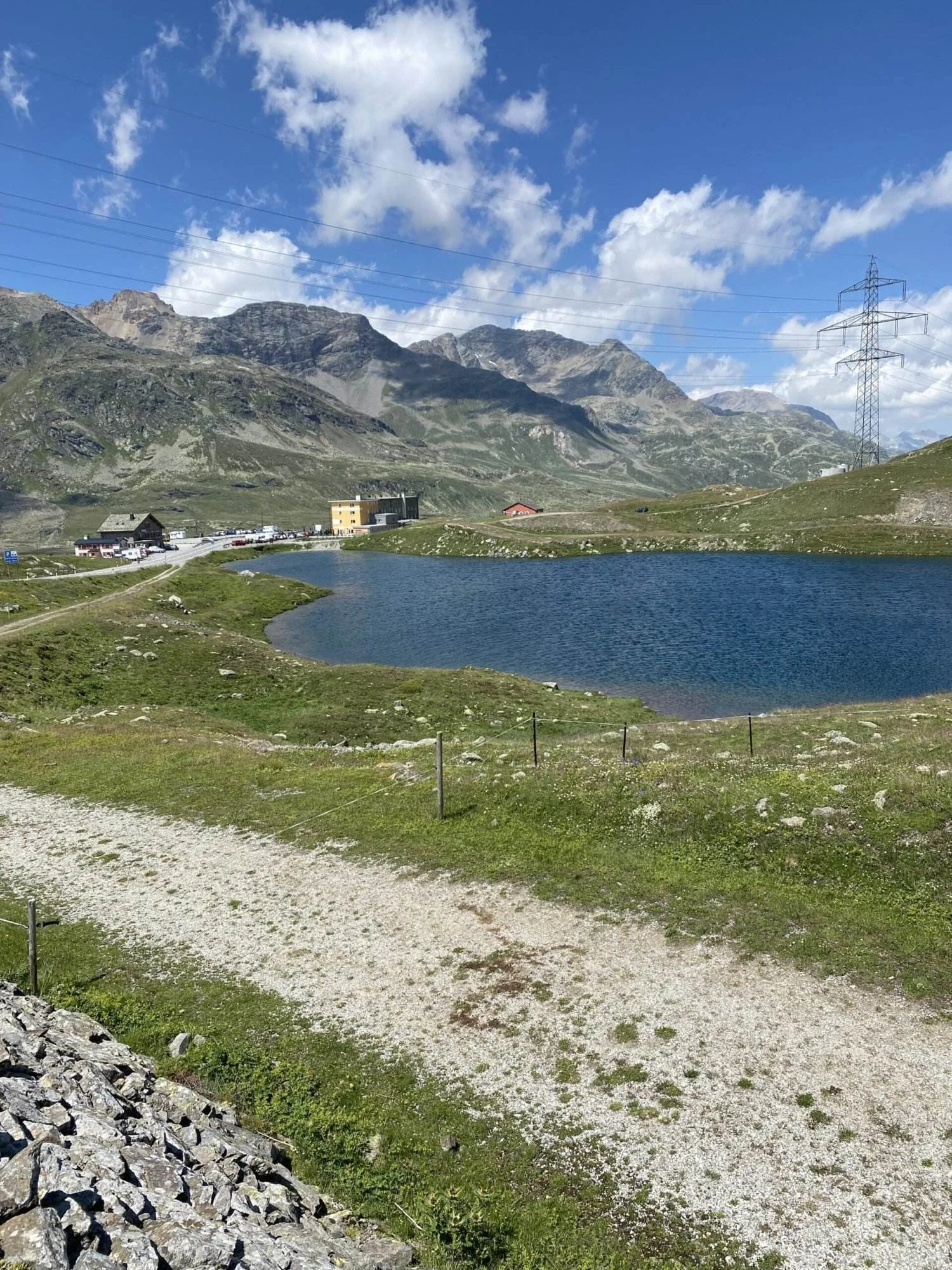Kleiner Bergsee neben einer Passstraße, mit Gebäuden am Ufer und hohen Strommasten vor einer alpinen Bergkulisse.