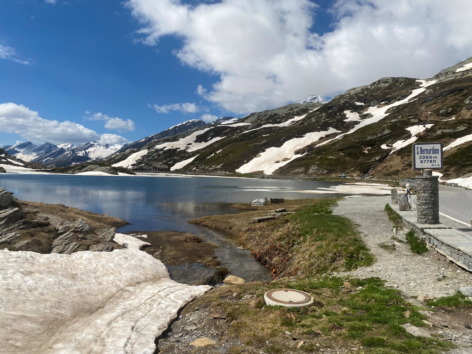 Bergsee am San-Bernardino-Pass mit teilweisem Schnee, ruhigem Wasser und umliegenden Bergen unter blauem Himmel, Weg und Passschild im Vordergrund.