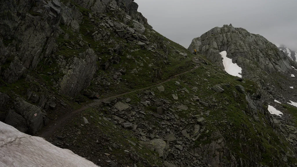 Bergpfad am Lago Sfundau, umgeben von Felsen und Schnee. Wanderer auf dem Weg.