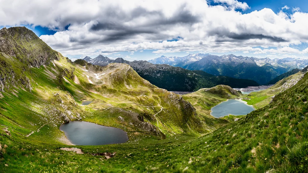 Bergpanorama mit dem Lago di Tom und grünen Wiesen unter bewölktem Himmel.