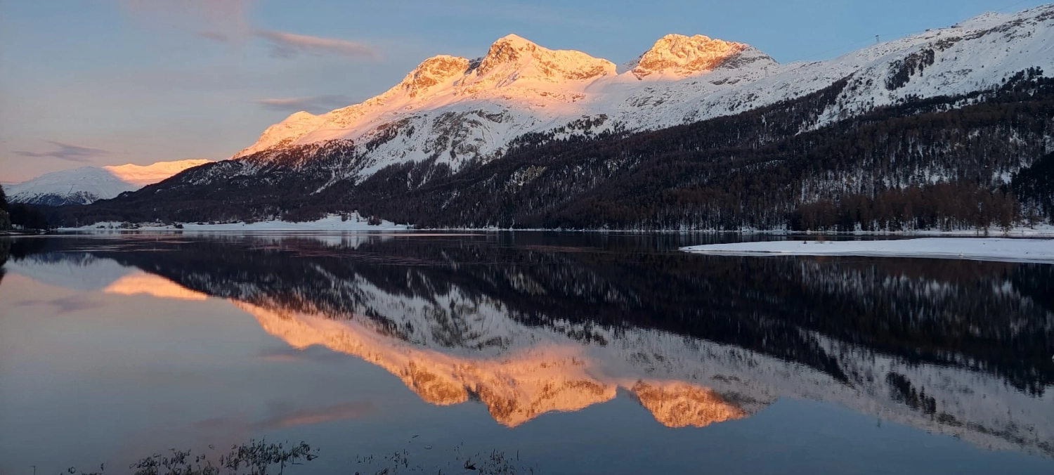 Silsersee im Winter: Verschneite Berge spiegeln sich im See bei Sonnenaufgang.