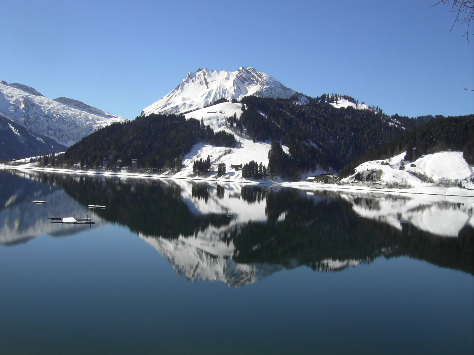 Wägitalersee-Spiegelung: Verschneiter Berg und dunkler Wald im klaren Wasser.