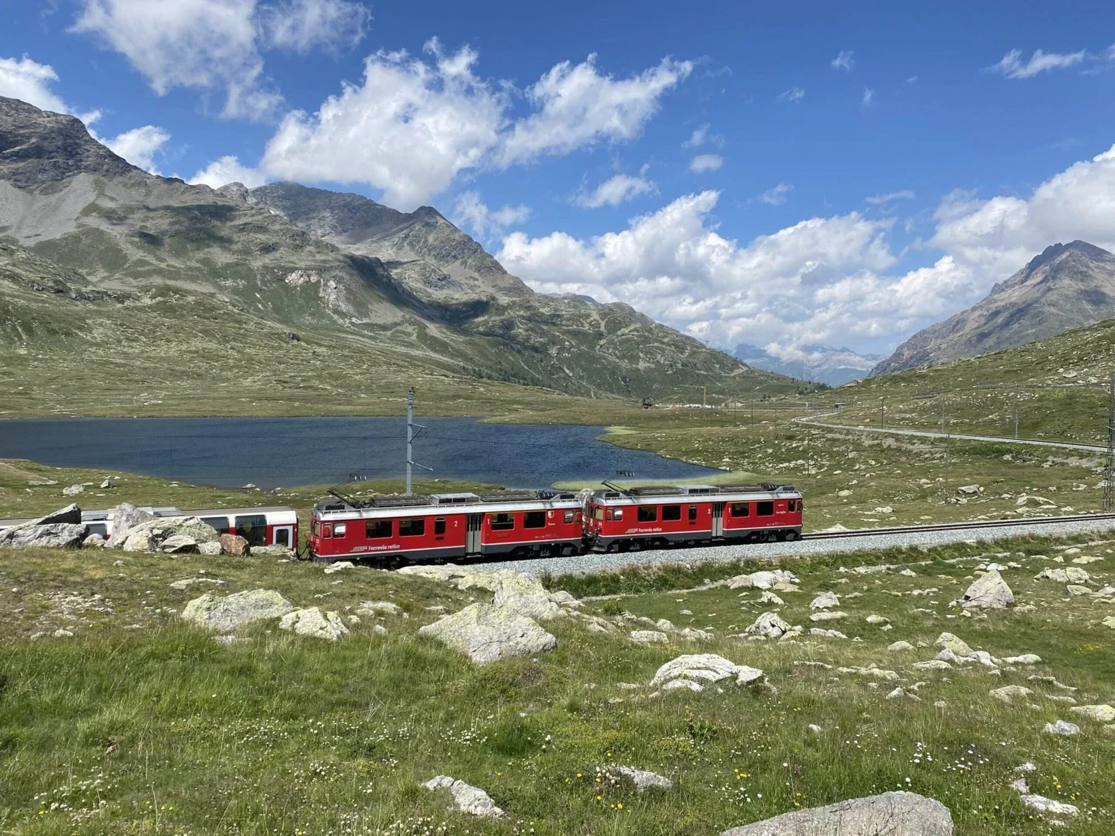 Roter Zug fährt durch alpine Hochebene am Bergsee vorbei, umgeben von Bergen, Wiesen und vereinzelten Felsen.